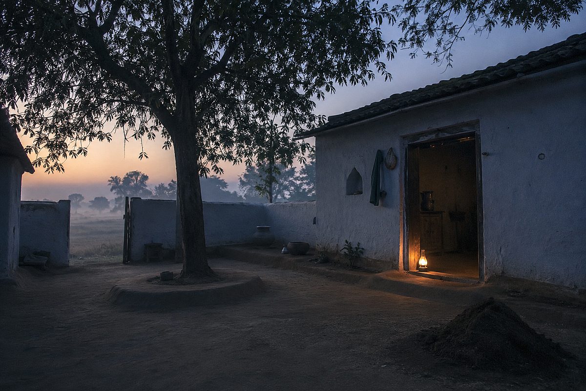 A farmhouse courtyard at first light, a single lamp burning in an open doorway